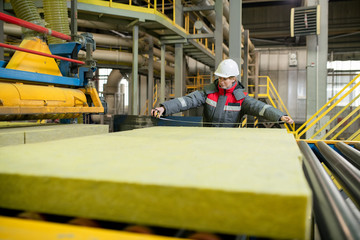 Production line worker in hardhat standing at conveyor belt and measuring construction material at...