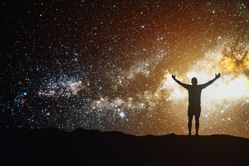 Night sky with stars cosmos and silhouette of a standing man with raised hands on the mountain....