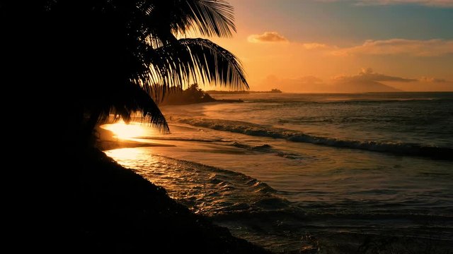 Beach under orange sunset light with silhouettes in the background. Waves break on the sand, aerial view, Papeete French Polynesia