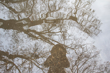 Reflection of a girl in a jacket with a hood in a puddle of water with trees on the background. A...
