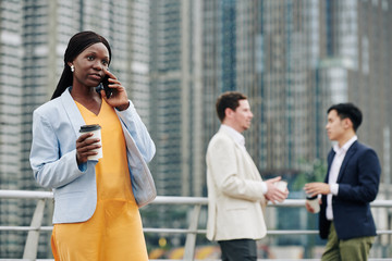 Pretty young Black businesswoman drinking take out coffee and talking on phone with business partner or assistant