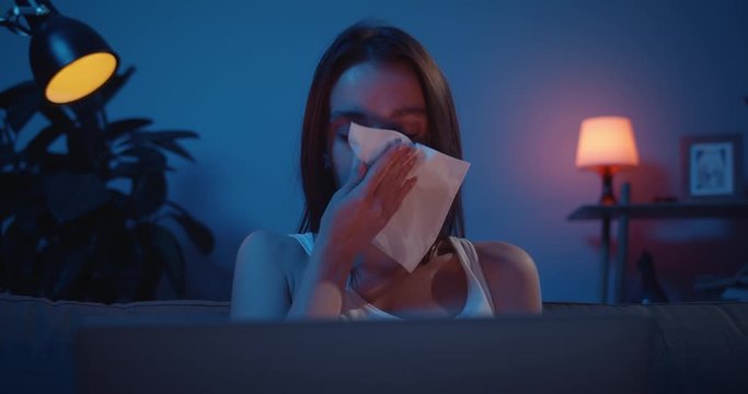 Sad millennial girl crying hardly and using tissues while watching drama on laptop. Unhappy woman in tears sitting on sofa with personal computer and worrying about personal problems.