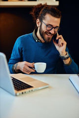 Young smiling caucasian bearded blogger sitting in his office, talking on the phone, holding cup of coffee and taking a break from work.