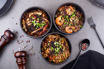 Wok noodles, with shrimp and other Asian ingredients, in a black plate decorated with peas, on a gray background . The concept of Asian cuisine.