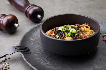 Wok noodles, with shrimp and other Asian ingredients, in a black plate decorated with peas, on a gray background . The concept of Asian cuisine.
