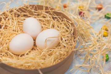 Three white eggs and colorful round sweets in a hay nest in a box. Top side view on a light blue wooden background. Copyspace for the wording. Easter holidays concept.