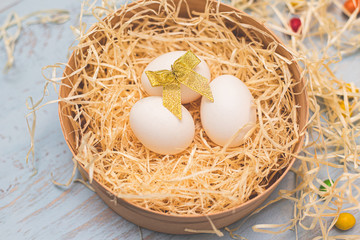 Three white eggs with a golden bow and colorful round sweets in a hay nest in a box. Top side view on a light blue wooden background. Copyspace for the wording. Easter holidays concept.