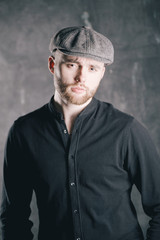 Serious handsome young bearded man in hat on grey background looking at camera. Portrait of man against grey wall.