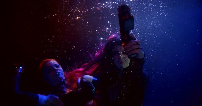 two girls with long hair float in the dark under the water. the woman in the foreground is aiming a gun, and the other one is floating up. blue, red and flickering white light