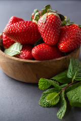 Top view of strawberries in wooden bowl and mint branch, with selective focus, on gray background in vertical