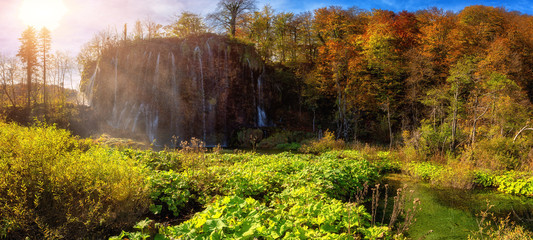Famous waterfall Prstavac in Plitvice lakes (Plitvicka jezera) national park, Croatia, large panorama. Amazing autumn sunny landscape with falling water, trees, sky and sun, outdoor travel background