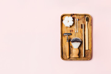 Zero waste concept. Natural bamboo toothbrush, bamboo ear sticks, razor, wooden tube, natural sponge, cotton flowers located on wooden tray on pink background. Flat lay, top view, copy space.