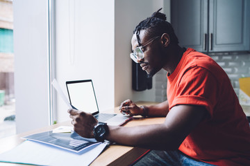 Concentrated young black businessman reading documents and using laptop in office