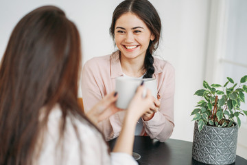 Young carefree brunette girls friends in casual with cups of tea meeting in bright cafe or kitchen...