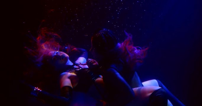 two girls with long hair float in the dark under the water. one of them hovers over the other, caressing her body and leg. close up. blue, red and flickering white light