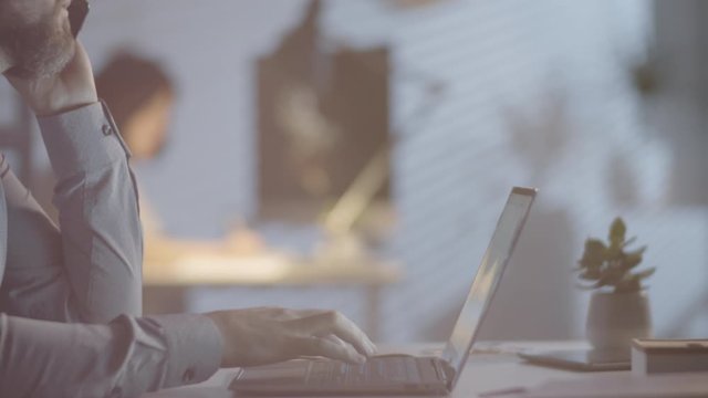 Tracking with slowmo of bearded man in glasses sitting at his desk and working on laptop while talking on mobile phone in dark office in evening