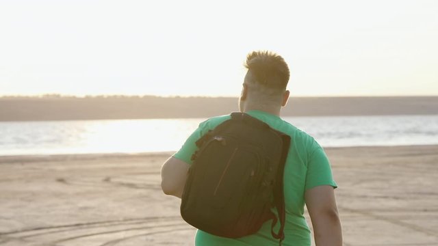 A plump guy in a green T-shirt goes to the water, throwing a backpack over his shoulder.