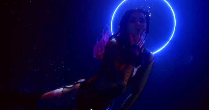 a brunette with long hair floats in the dark under the water. a white shimmering Hoop forms a halo around her head. blue and red contour light