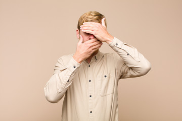 young blonde man covering face with both hands saying no to the camera! refusing pictures or...