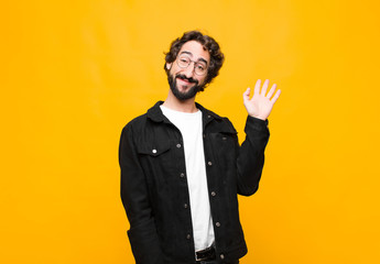 young crazy handsome man smiling happily and cheerfully, waving hand, welcoming and greeting you, or saying goodbye against orange wall