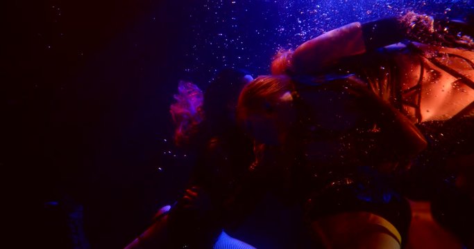 Three Girls Float In The Dark Under The Water. In The Foreground, A Woman In High Boots Floats By. The Other Two Are Kissing In The Background. Blue, Red And Flickering White Light