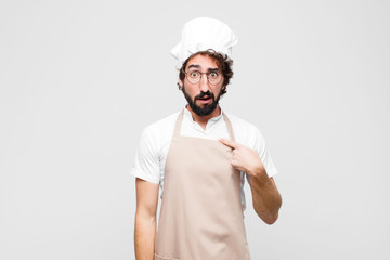 young crazy chef feeling confused, puzzled and insecure, pointing to self wondering and asking who,...