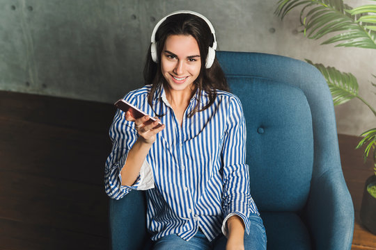 Hipster girl in earphones listening to music in online app sitting on sofa in her own apartment. Young lady downloading audio apps, digital tools and solutions on smartphone. Streaming.