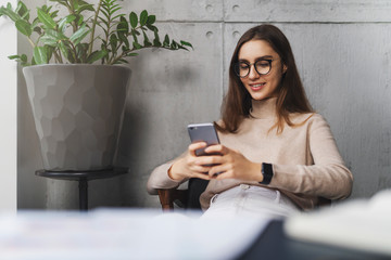 Young girl in glasses sitting in armchair and texting on smartphone. Entertaining and educating...