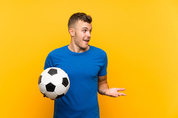 Young handsome blonde man holding a soccer ball over isolated yellow background with surprise...