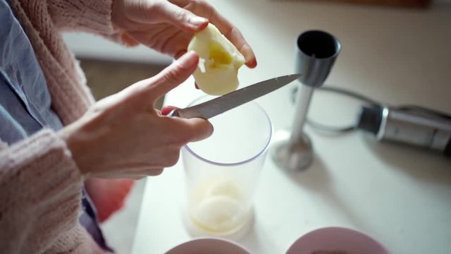 female hands with knife cutting apple for smoothie in transparent blender cup, slow motion