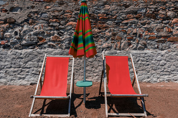 View of two inviting empty orange wooden chairs for sunbathing and a table with ditto parasol, standing in the sand on the beach in Italy, as a background a stone wall with large yellow boulders.