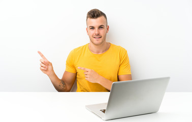Young blonde man in a table with a laptop pointing finger to the side