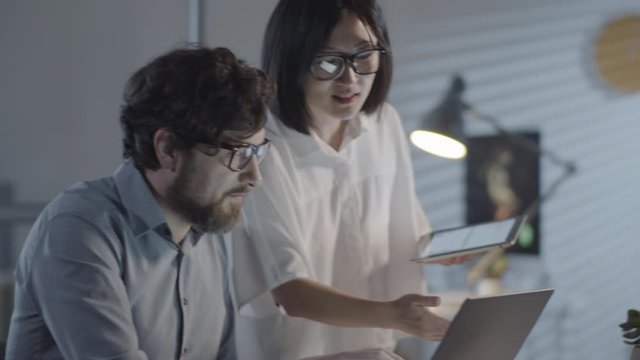 Slowmo tracking of cheerful Asian businesswoman with tablet and bearded man with laptop discussing project they are working on in dark office in evening