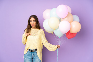 Young woman catching many balloons over isolated on purple background with surprise facial...