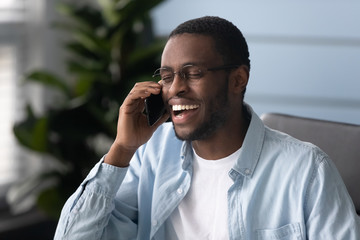 African ethnicity guy laughing during smartphone conversation seated indoors