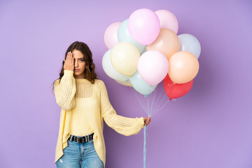 Young woman catching many balloons over isolated on purple background covering a eye by hand