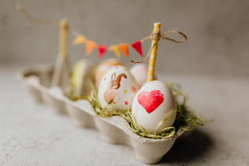 Ostern - Mit Aquarellefarben bemalte Ostereier auf Ostergras in Eierkarton mit Girlande auf hellen...