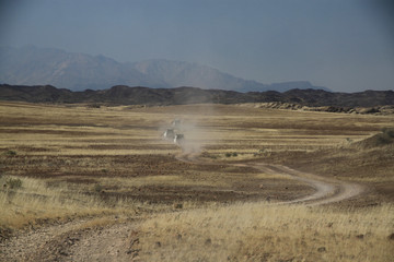 samochody terenowe w oddali na polnej drodze pomiędzy wyschniętymi trawami afrykańskiej sawanny z górami w tle