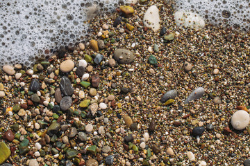 Closeup top view of texture of sandy beach with colourful small rounded pebbles and splashing foamy sea waves. Abstract organic natural photo background.