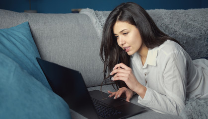 Young pretty concentrated lady holding eyewear in her hand reading from laptop while lying on her abdomen on sofa in flat