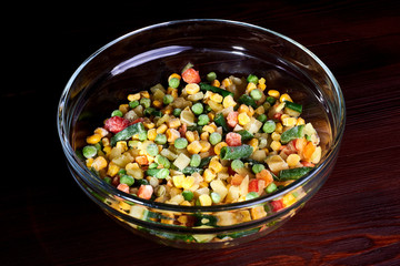 Frozen different vegetables in a glass plate on a wooden background.