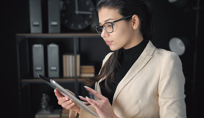 Focused businesswoman wearing spectacles using electronic tab in dark office background