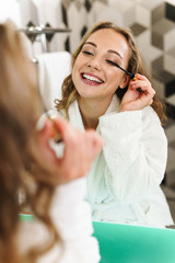 Image of young woman in housecoat applying makeup after shower in bathroom