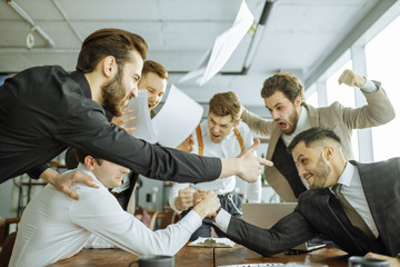 cheerful colleagues enjoy free, leisure time at work place, two male playing arm wrestling in office, co-workers support them