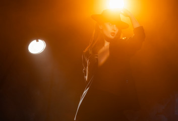 Fototapeta na wymiar Beautiful braless actress girl, wearing an unbuttoned blazer and a hat, poses and dances in the warm light rays of scenic illuminators in the theatrical fog. Noir, lifestyle and trendy design.