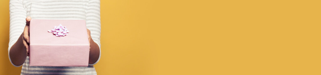 Portrait of young beautiful african american woman holding pink gift box on bright yellow background