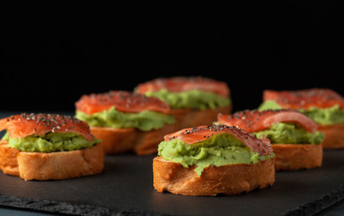 Salmon bruschettas with avocado on a black background, decorated with chia seeds.
