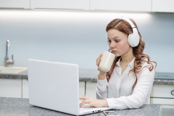 Teen girl barks at home in the kitchen on a laptop, while drinking coffee, looks at the laptop screen, next to a smartphone