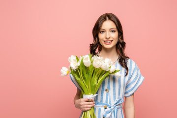 cheerful girl smiling at camera while holding bouquet of white tulips isolated on pink