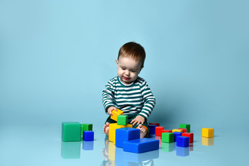 Obraz premium Little baby boy in stylish casual clothing barefoot sitting on floor and playing with colorful toy cubes over blue wall background